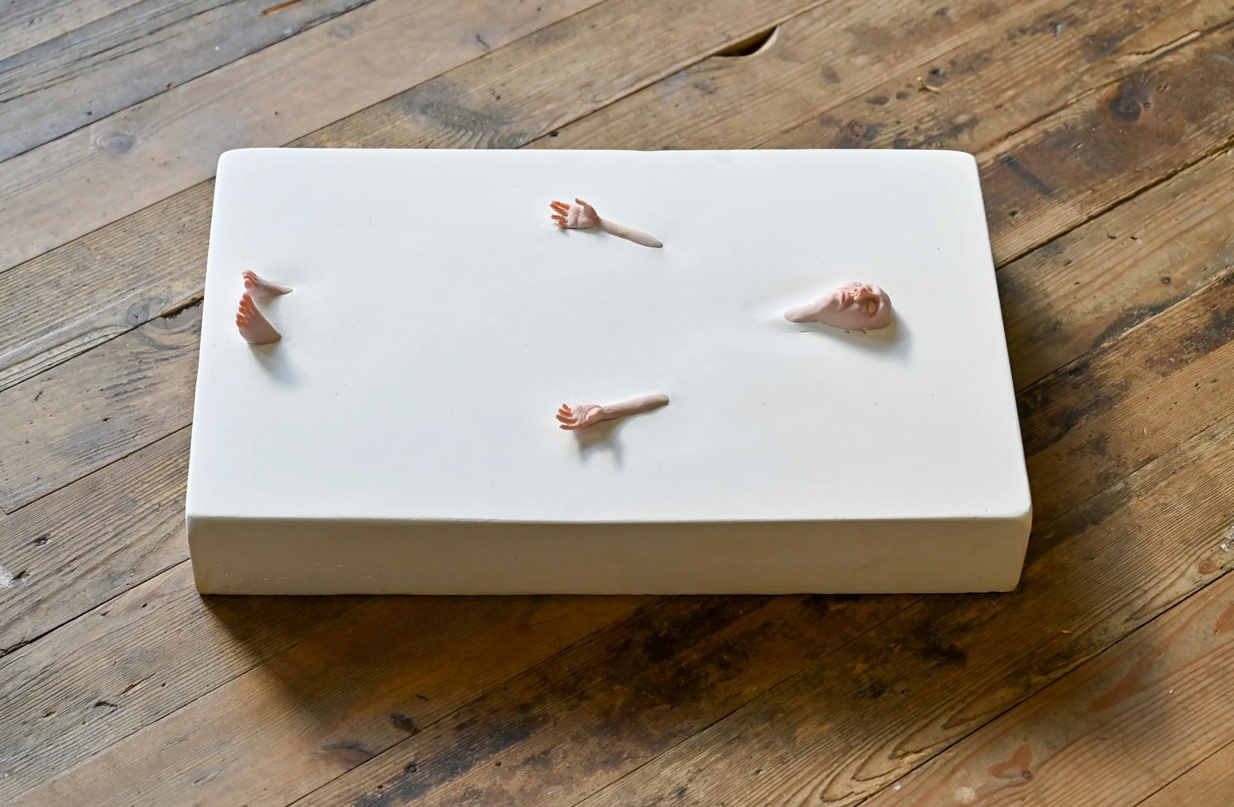 A white rectangular box on a wooden floor with small sculpted human body parts, including two hands, two feet, and a head, arranged on top of it.