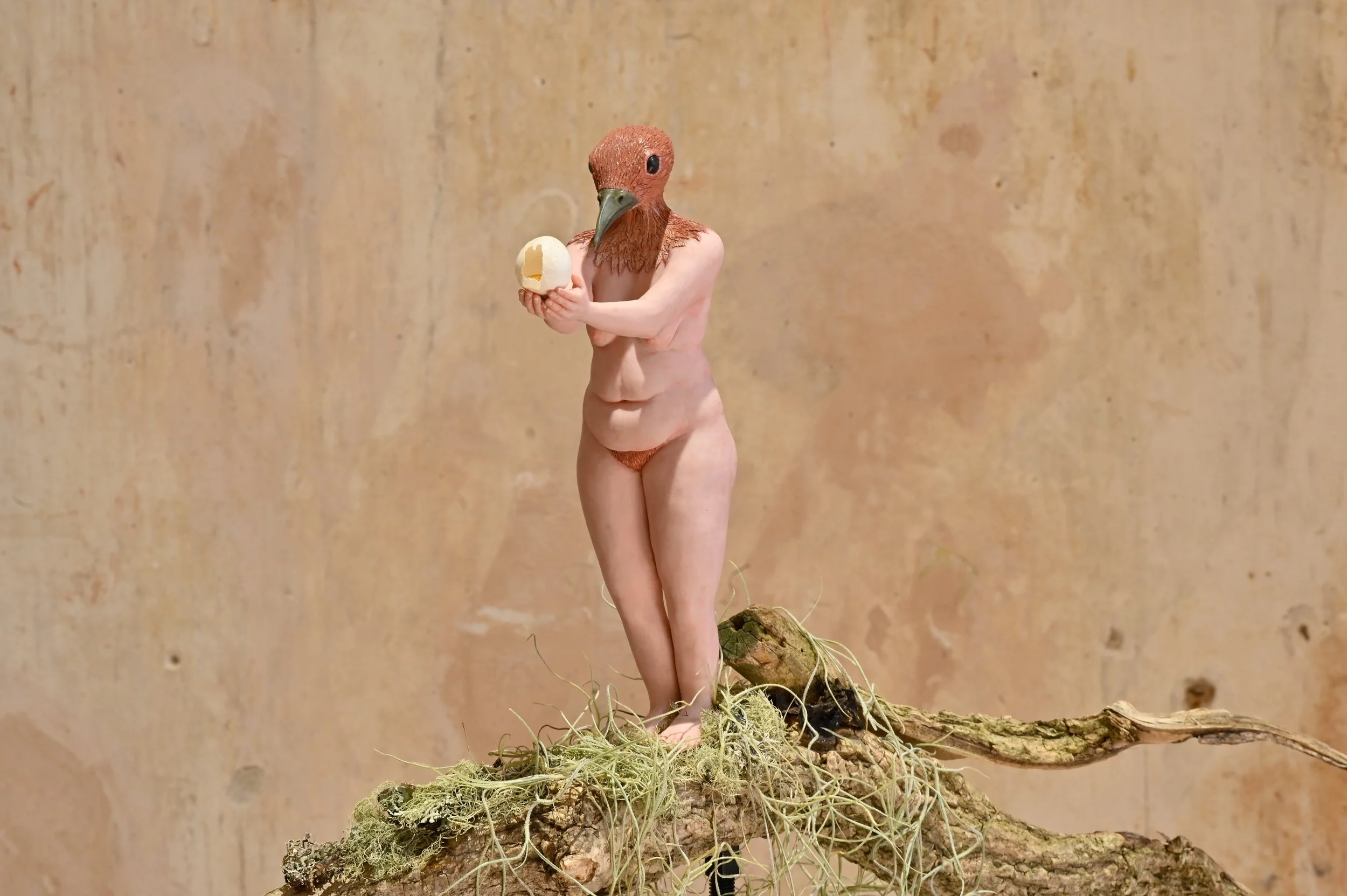 A humanoid figure with a bird's head and human body holding an eggshell, standing on a piece of wood with moss and lichen, against a textured beige background.