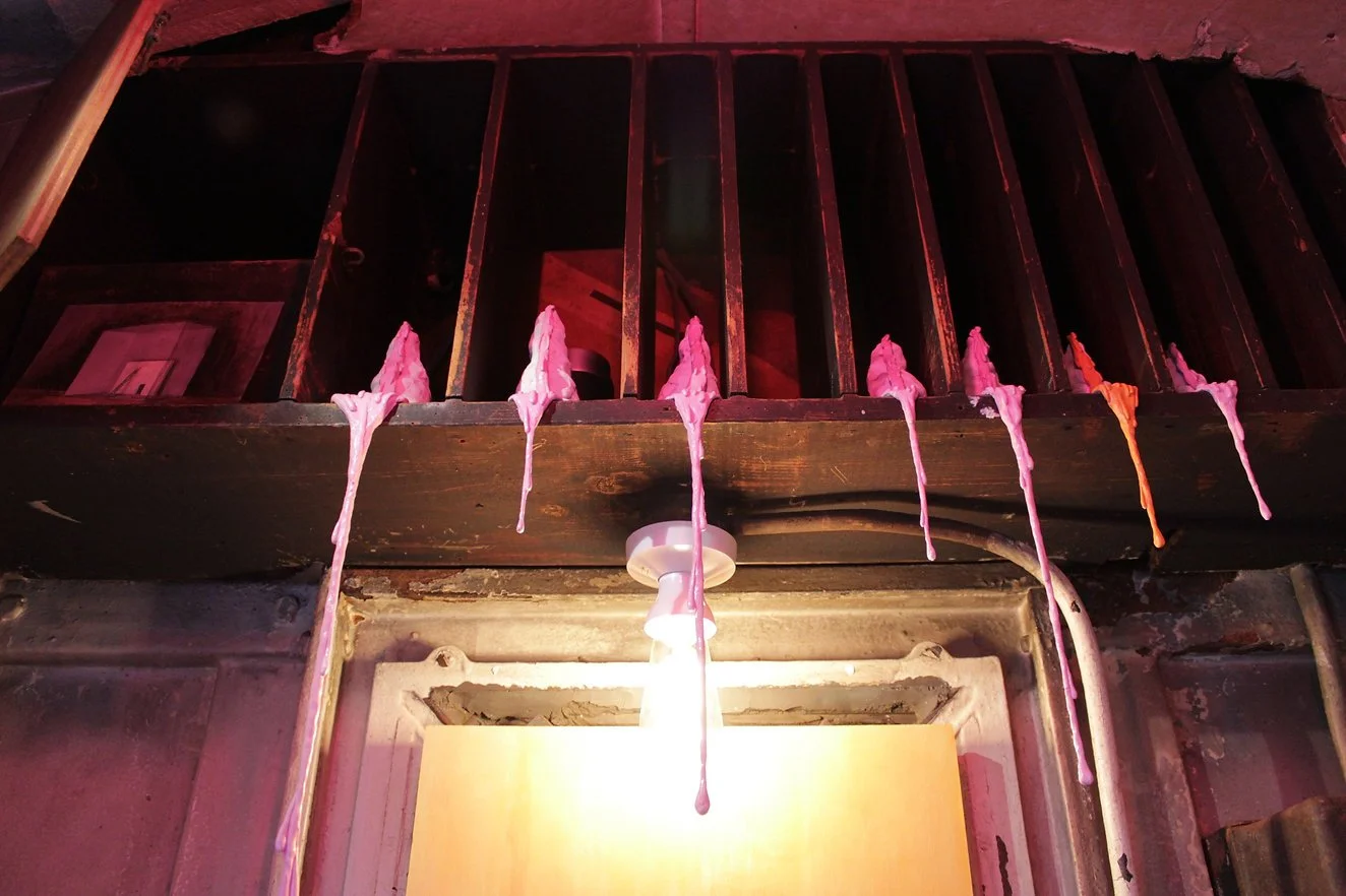Pink and orange melted candle wax dripping from a metal candlestick holder onto a wooden shelf with vertical bars above. The scene is dimly lit and has a rustic, somewhat worn appearance.