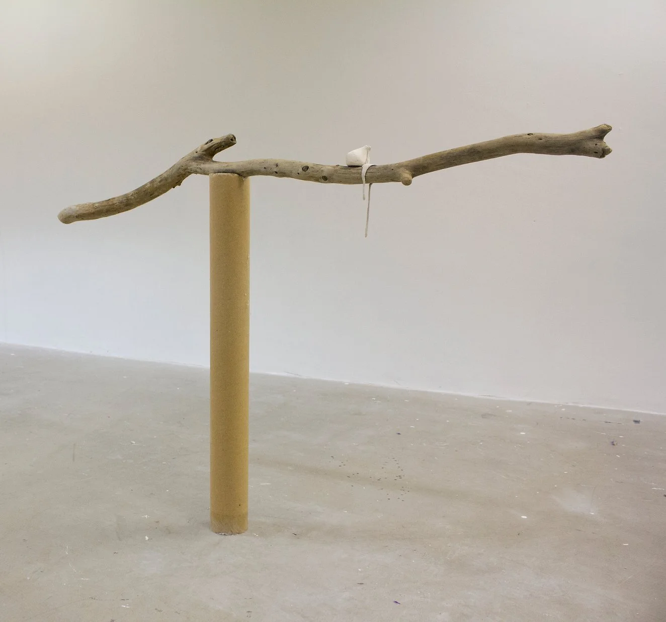 A minimalist sculpture featuring a horizontal driftwood branch supported by a vertical cylindrical cardboard post against a plain white wall.