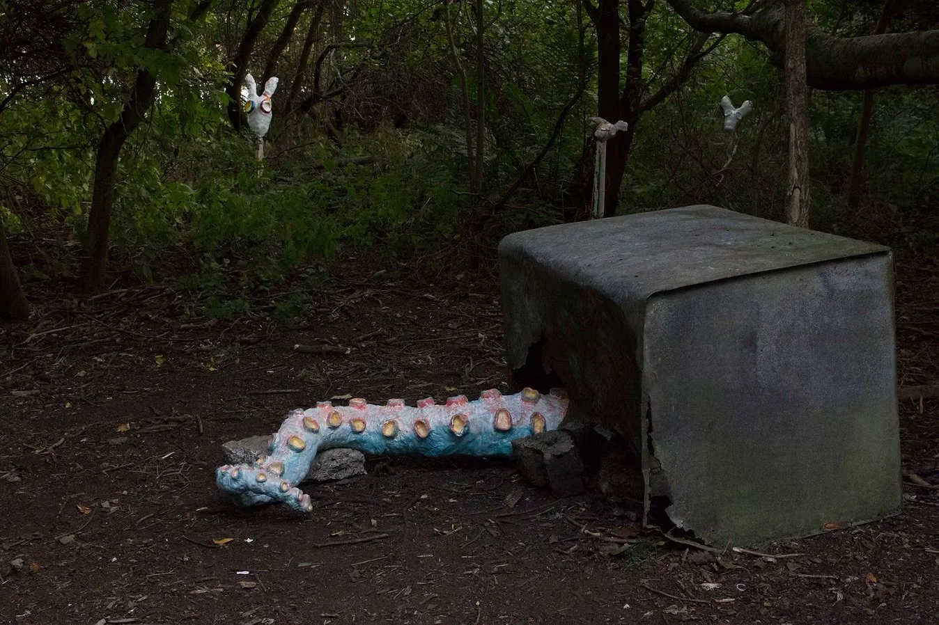 A surreal outdoor scene with a stuffed octopus reaching into a concrete box, and several animal masks hanging from tree branches in a wooded area.