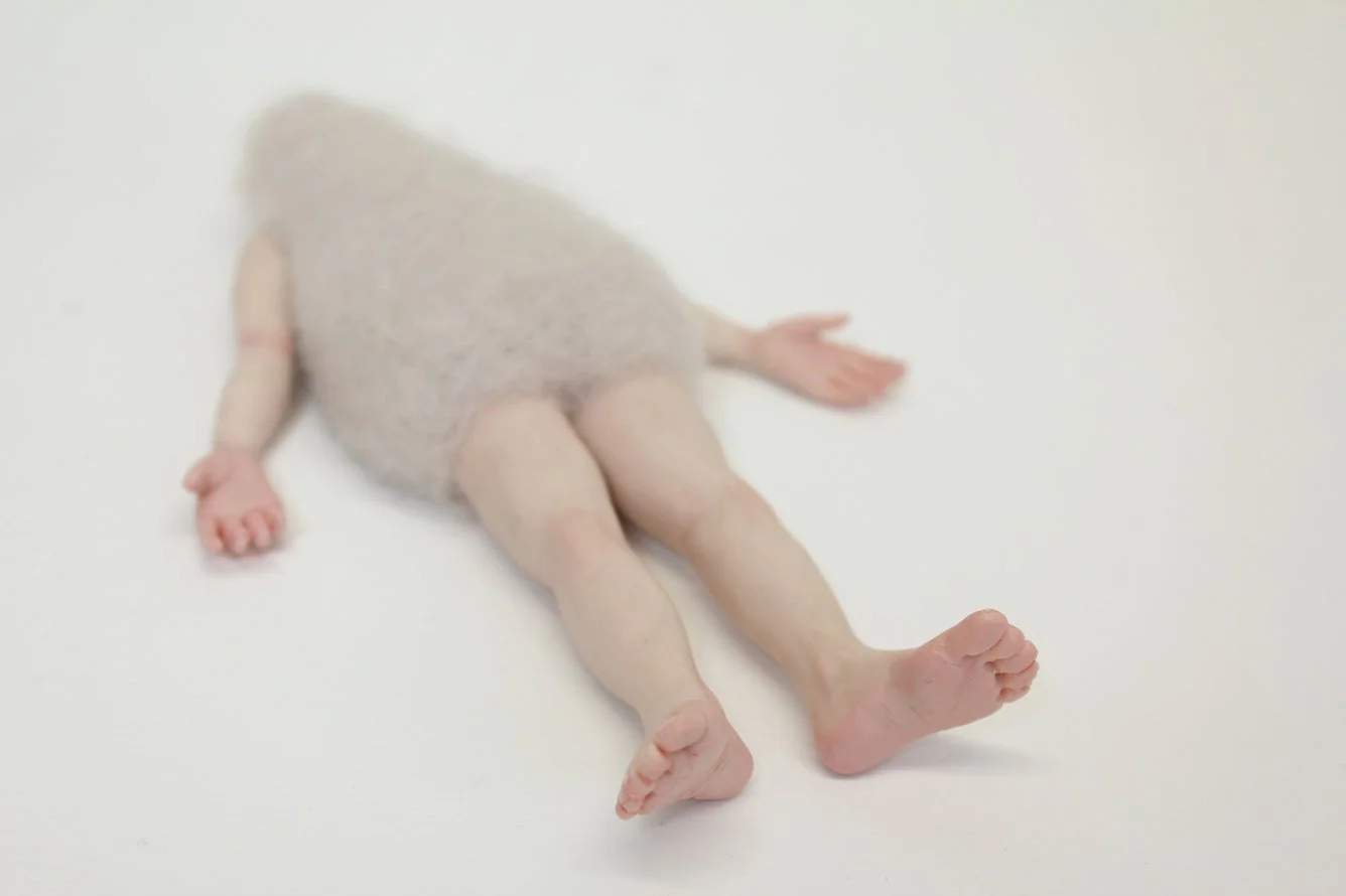 A surreal sculpture of a person with a head covered in a fluffy white material, lying down with arms and legs slightly splayed against a plain white background.