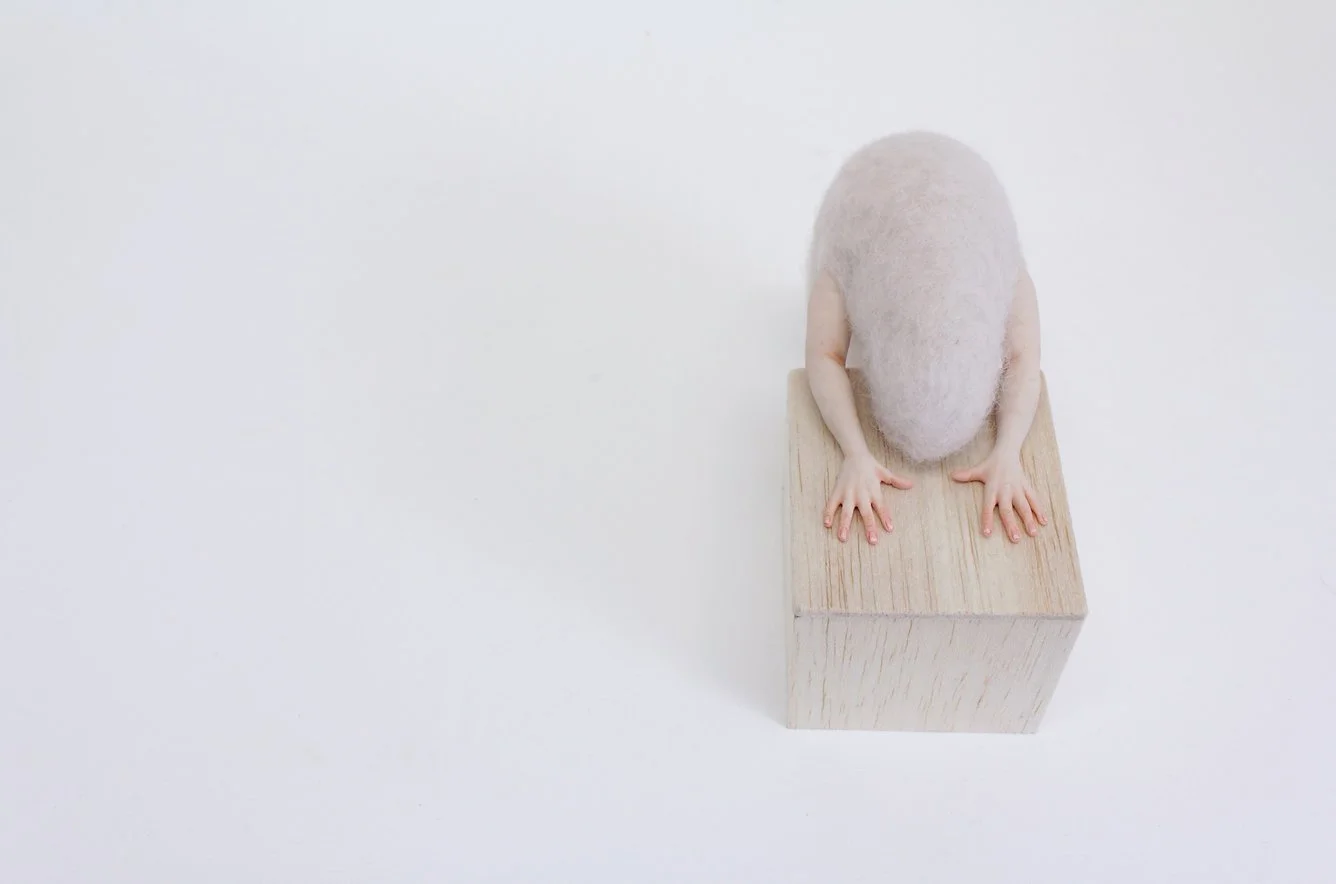 A small figure with a fuzzy, light-colored head, yellow arms, and pink hands, sitting on a wooden block against a plain white background, facing downward.