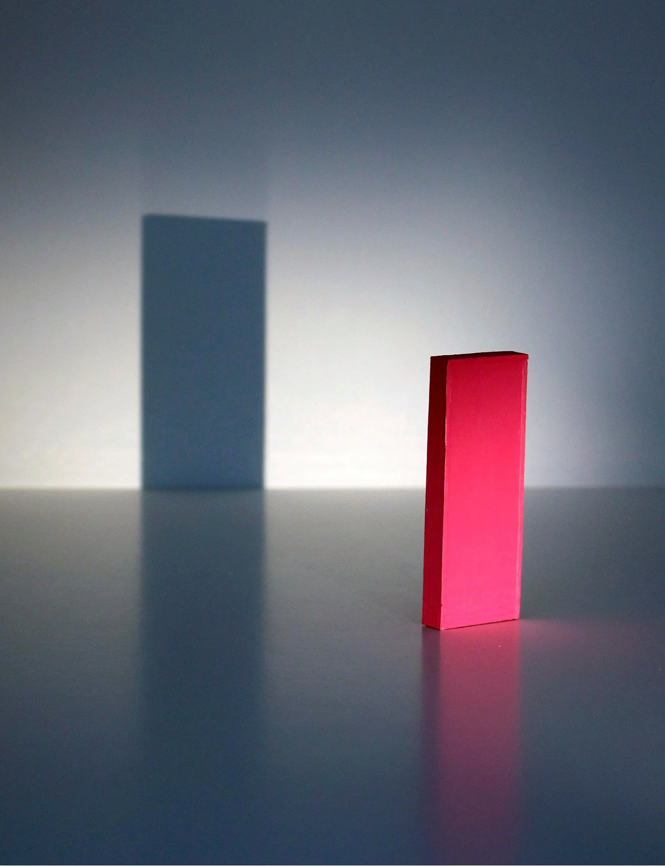 A red rectangular block standing upright on a flat surface with a shadow cast on the wall behind it. A blue rectangular block in the background has a larger shadow on the wall.