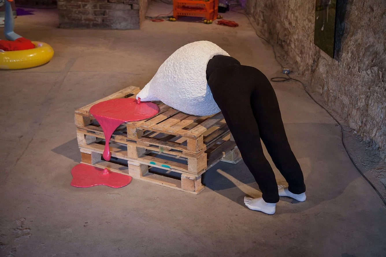 An art installation featuring a person with a white textured conical object over their head. The person is leaning forward with their hands and feet on the ground, wearing black pants and white socks. The cone is placed on a wooden pallet, with red p