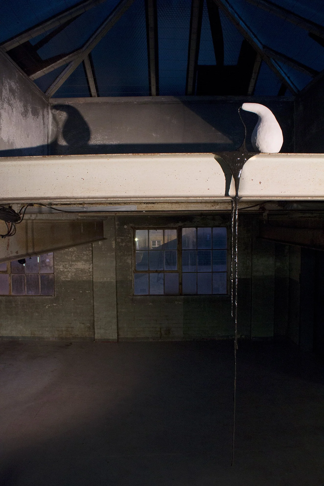 A white ceramic pot sitting on a ledge with black liquid dripping from it, inside a dimly lit industrial space with brick walls and large windowpanes