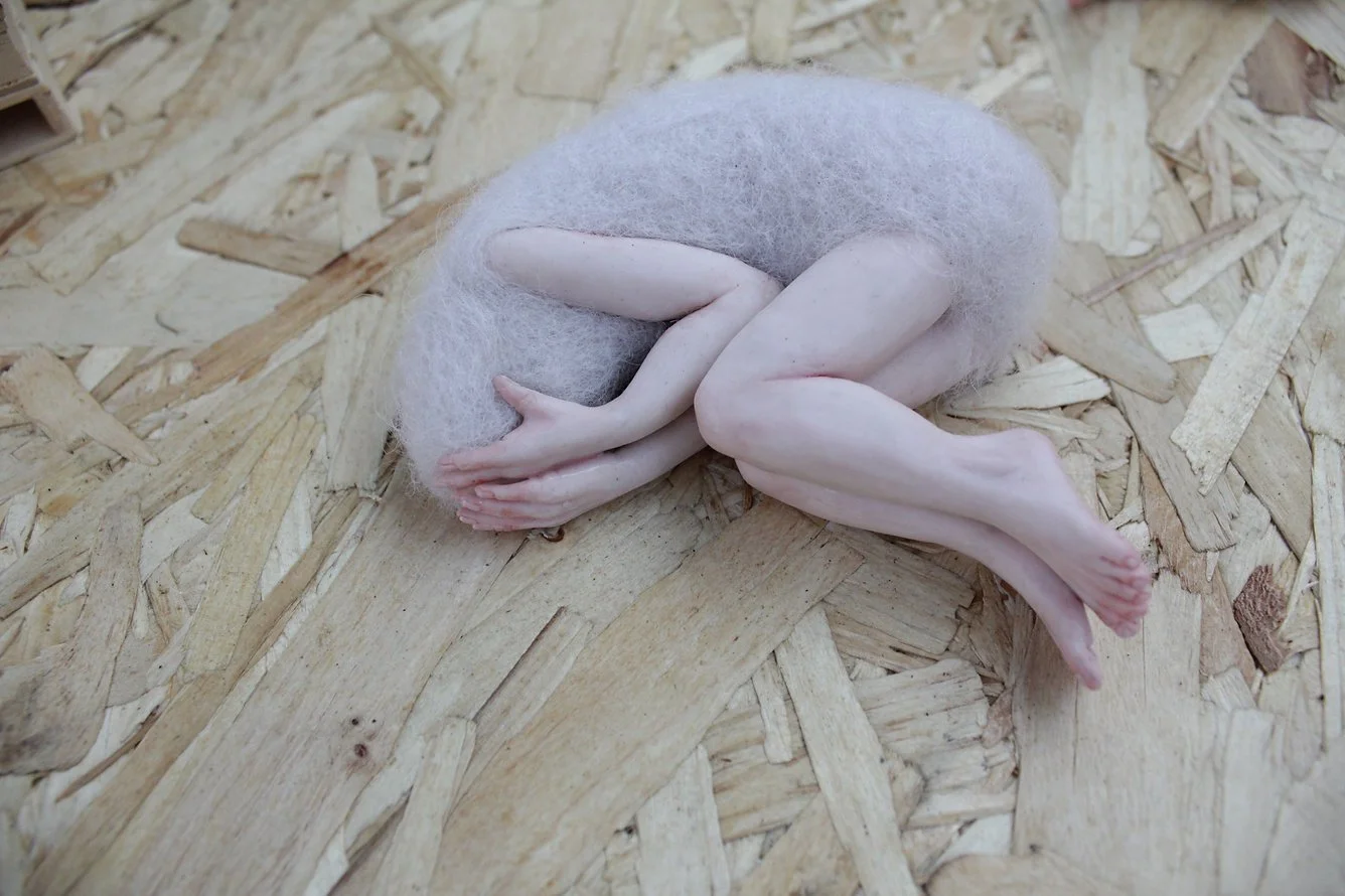 A  fragile felted figurine, curled up in a fetal position, on a wooden surface.