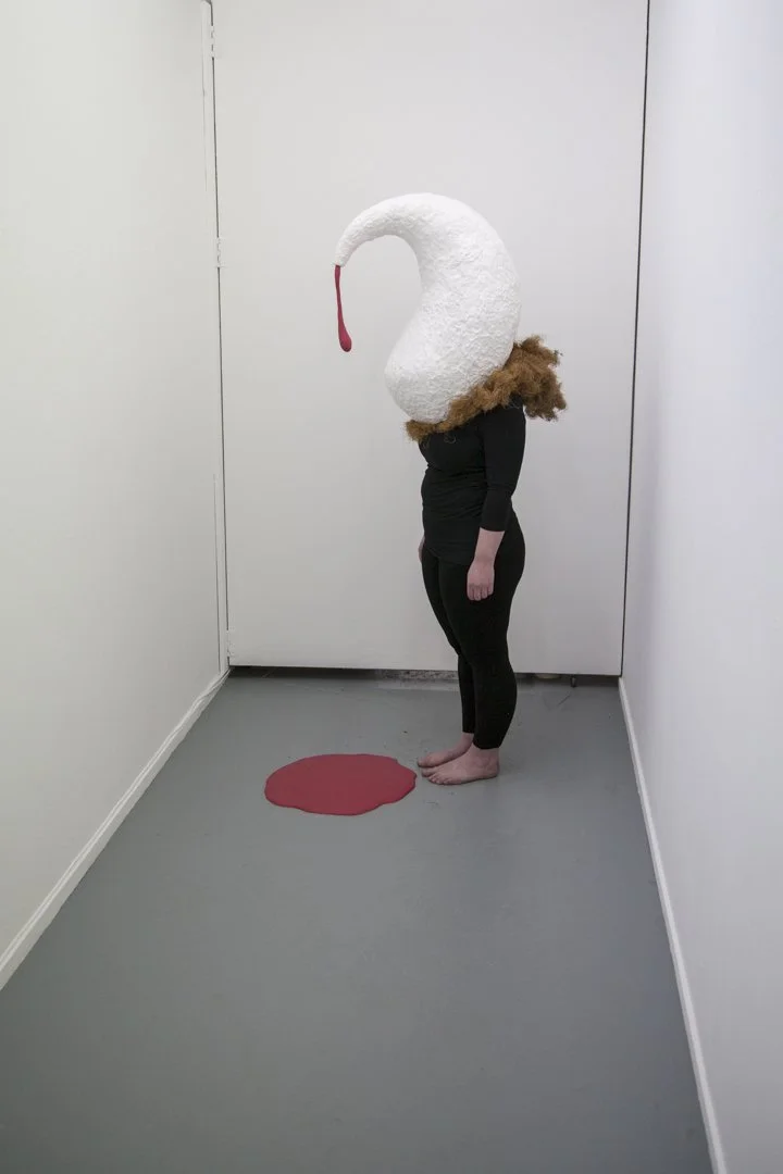 Person standing in a narrow room wearing a swan-like head mask, with a red spot painted on the floor in front of them.