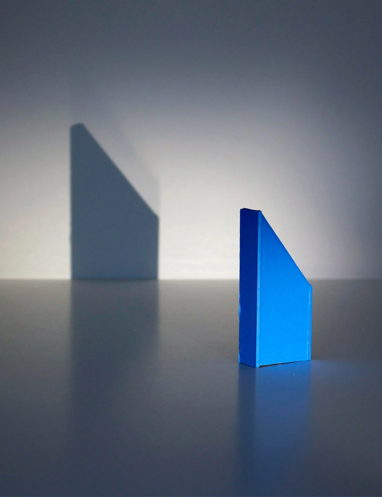 A blue geometric prism casting a dark blue shadow on a white surface with a gradient gray background.