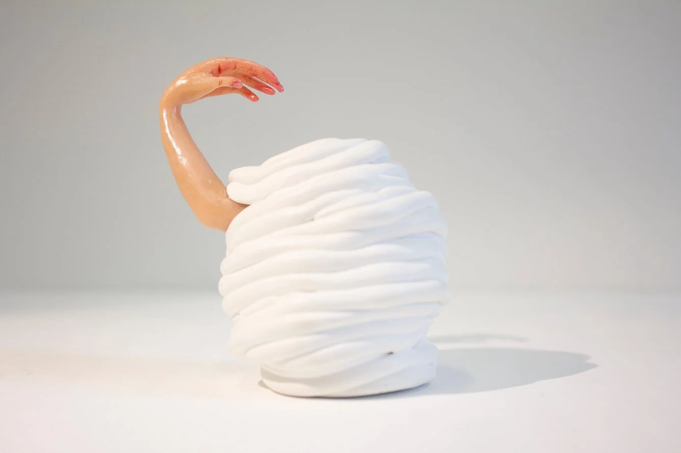 Whimsical sculpture of a a body made of white meringue-like with an emerging hand, on a plain surface against a light background.