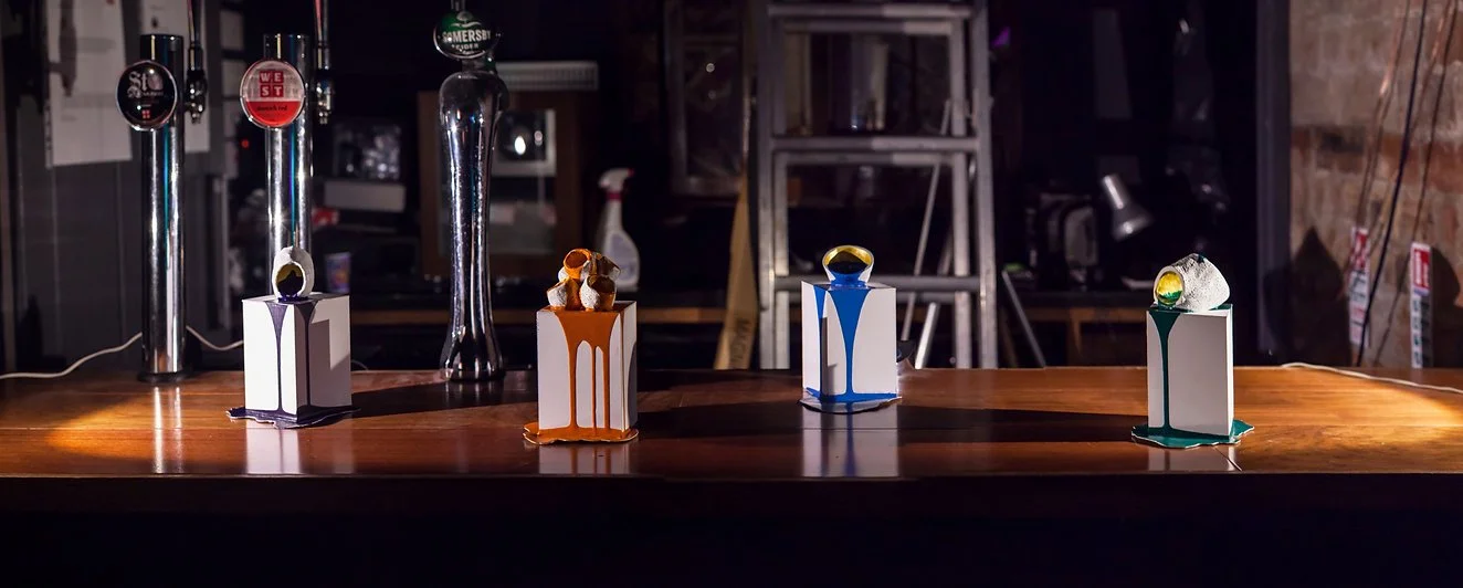 Four beer taps and four white boxes with colorful drips, each topped with a ring-shaped object, arranged on a wooden bar counter.