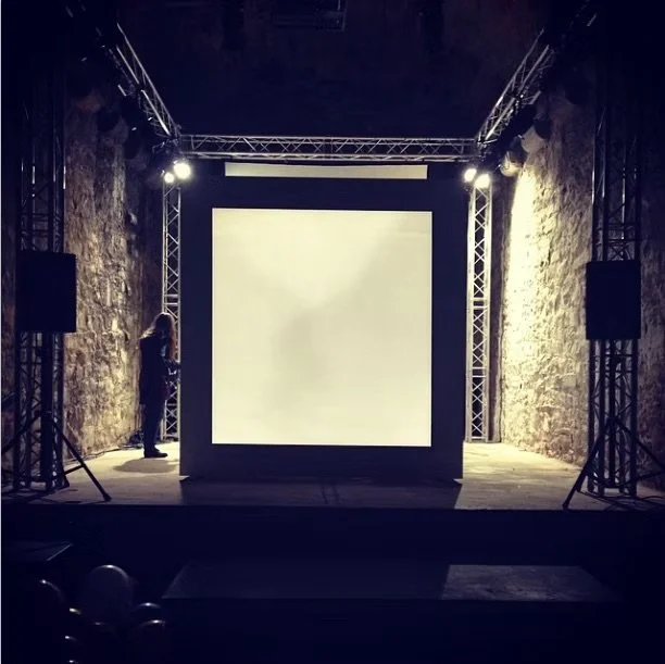 An indoor stage setup with a large blank screen and two side speakers, surrounded by stone walls and lighting equipment.