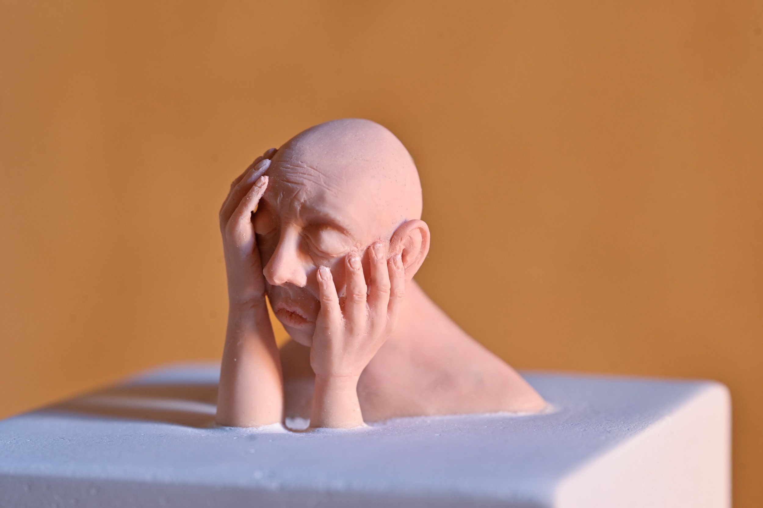 A sculpture of a person with a bald head, closed eyes, and hands covering parts of their face, showing a distressed or contemplative expression, set against a plain background.