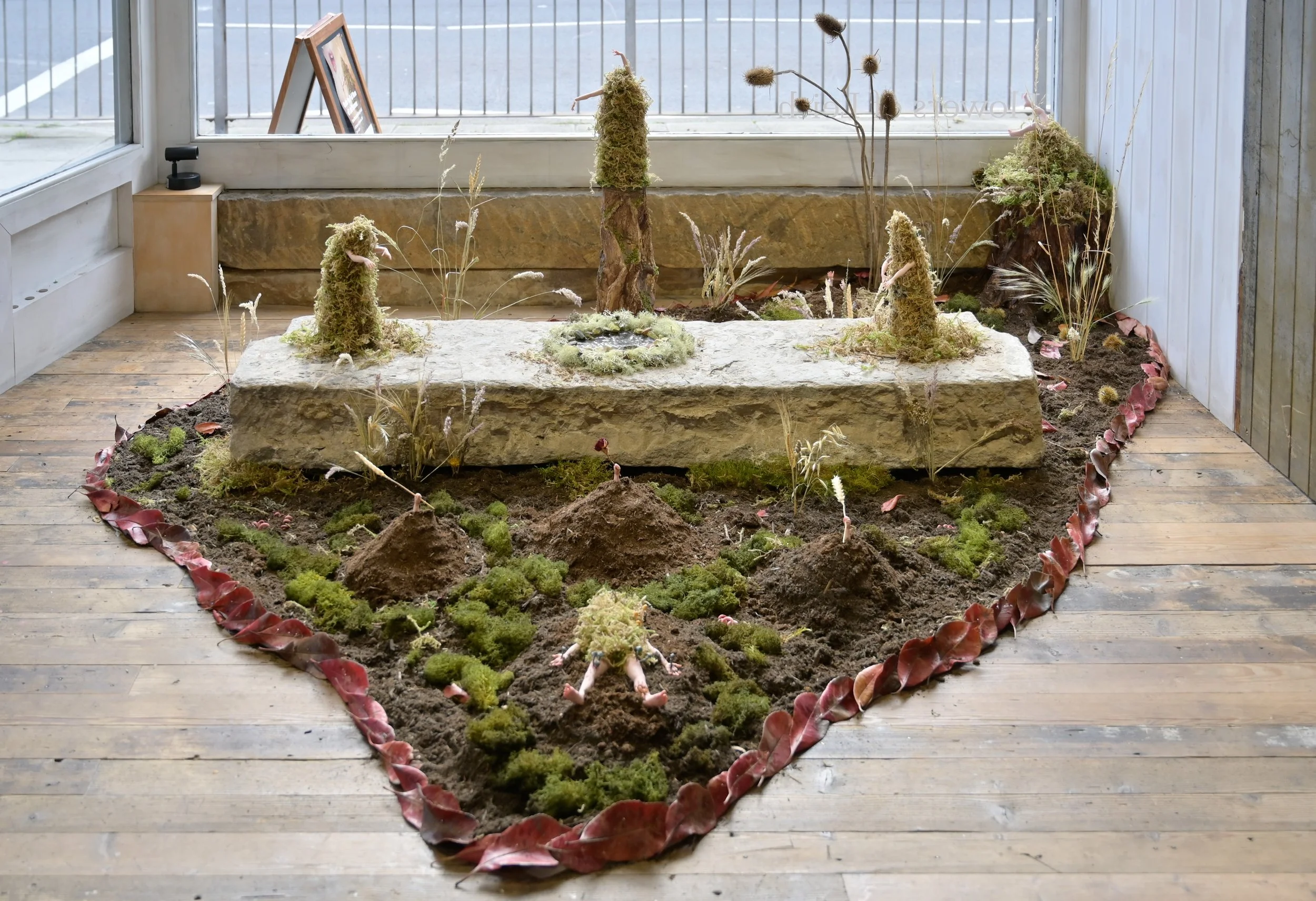Indoor art installation display with moss, soil, and plant sculptures, surrounded by red leaves on a wooden floor, in front of a large window.