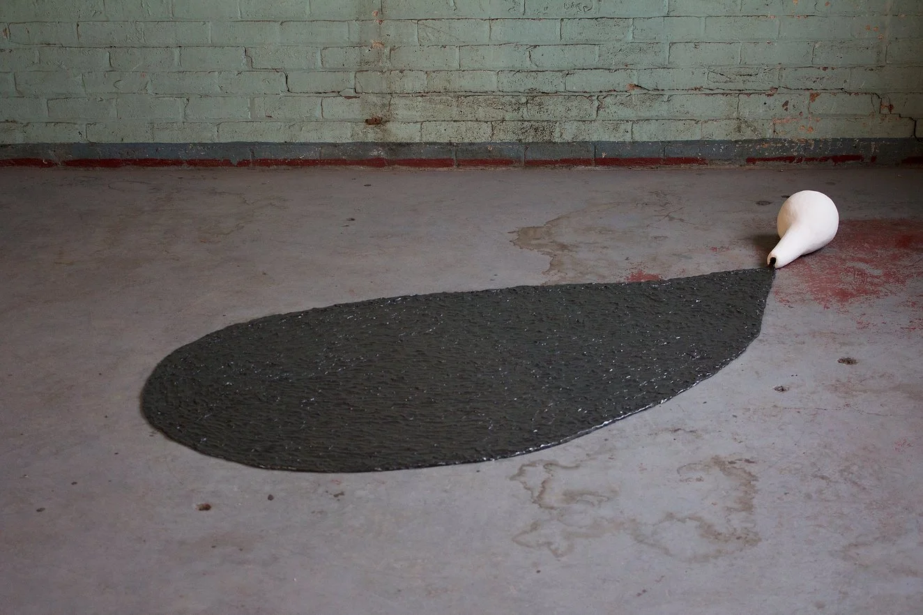 An art installation featuring a white ceramic pot lying on the floor with a black area painted underneath it, resembling a droplet or puddle, against a gray brick wall background.