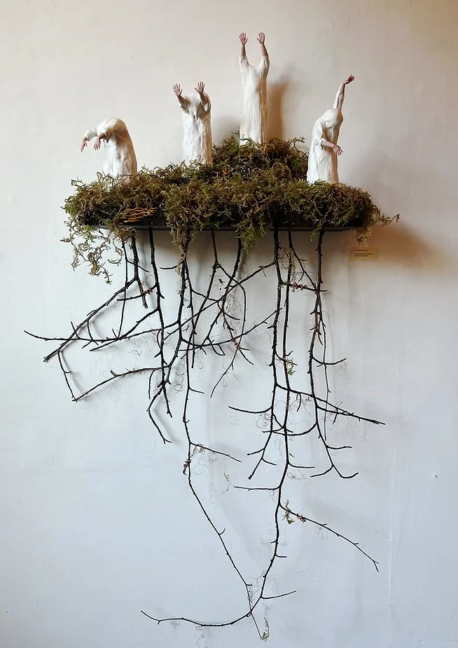 Art installation with four white plaster-covered figures with raised arms, positioned on a moss-covered platform with hanging branches resembling roots, mounted on a plain white wall.
