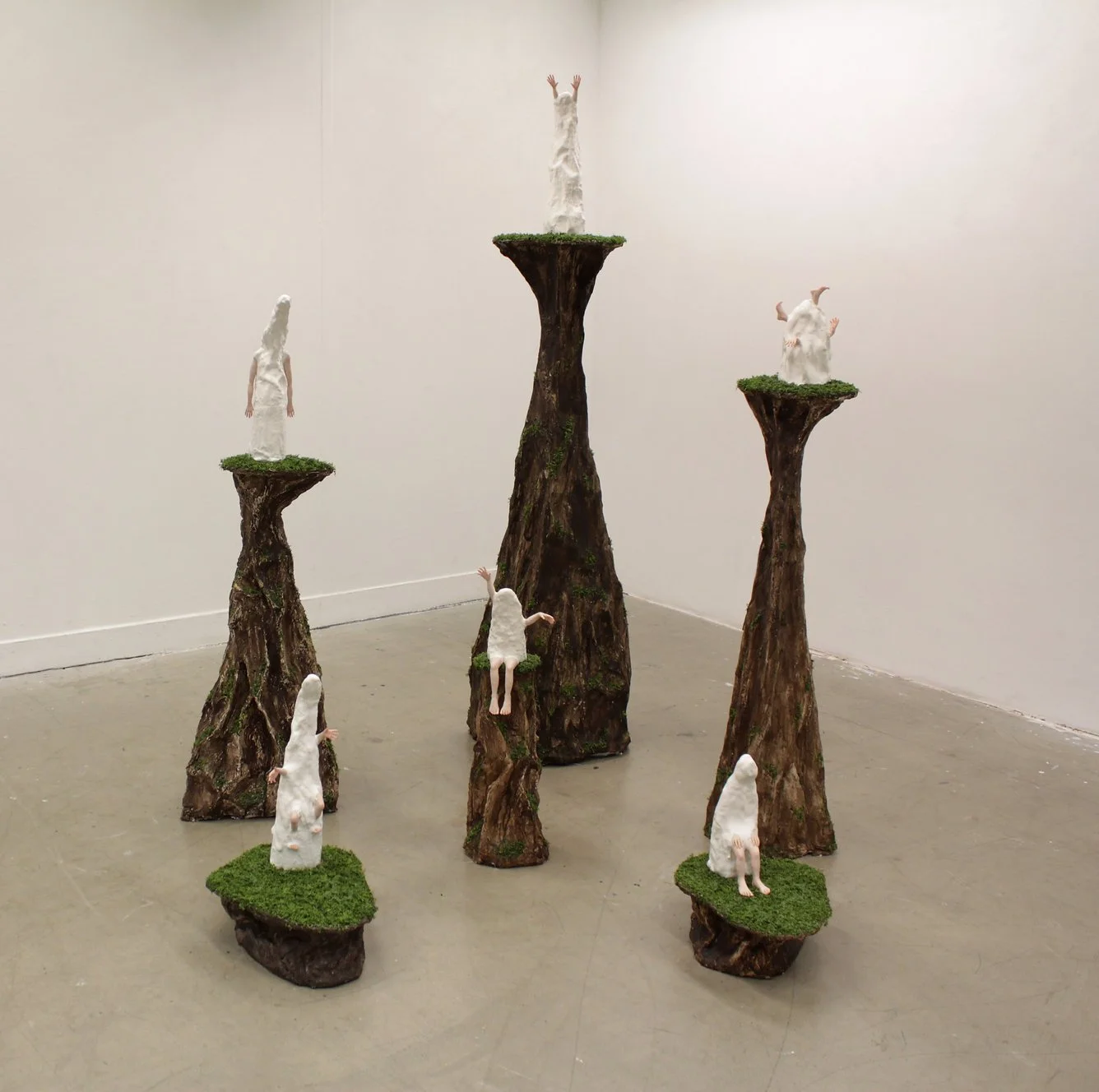 Art installation with wooden platforms resembling trees, topped with plant moss, and white sculptures of anthropomorphic creatures with animal features like horns and tails, arranged in a forest-like display.