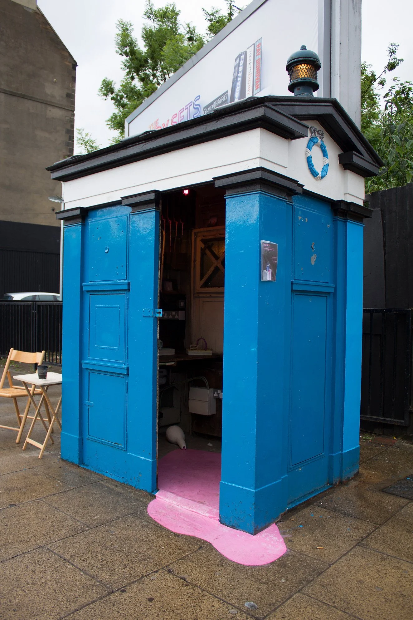 A small, blue, wooden phone booth with a pink rug on the ground inside, located outdoors on a rainy day, with a tree and billboard in the background.