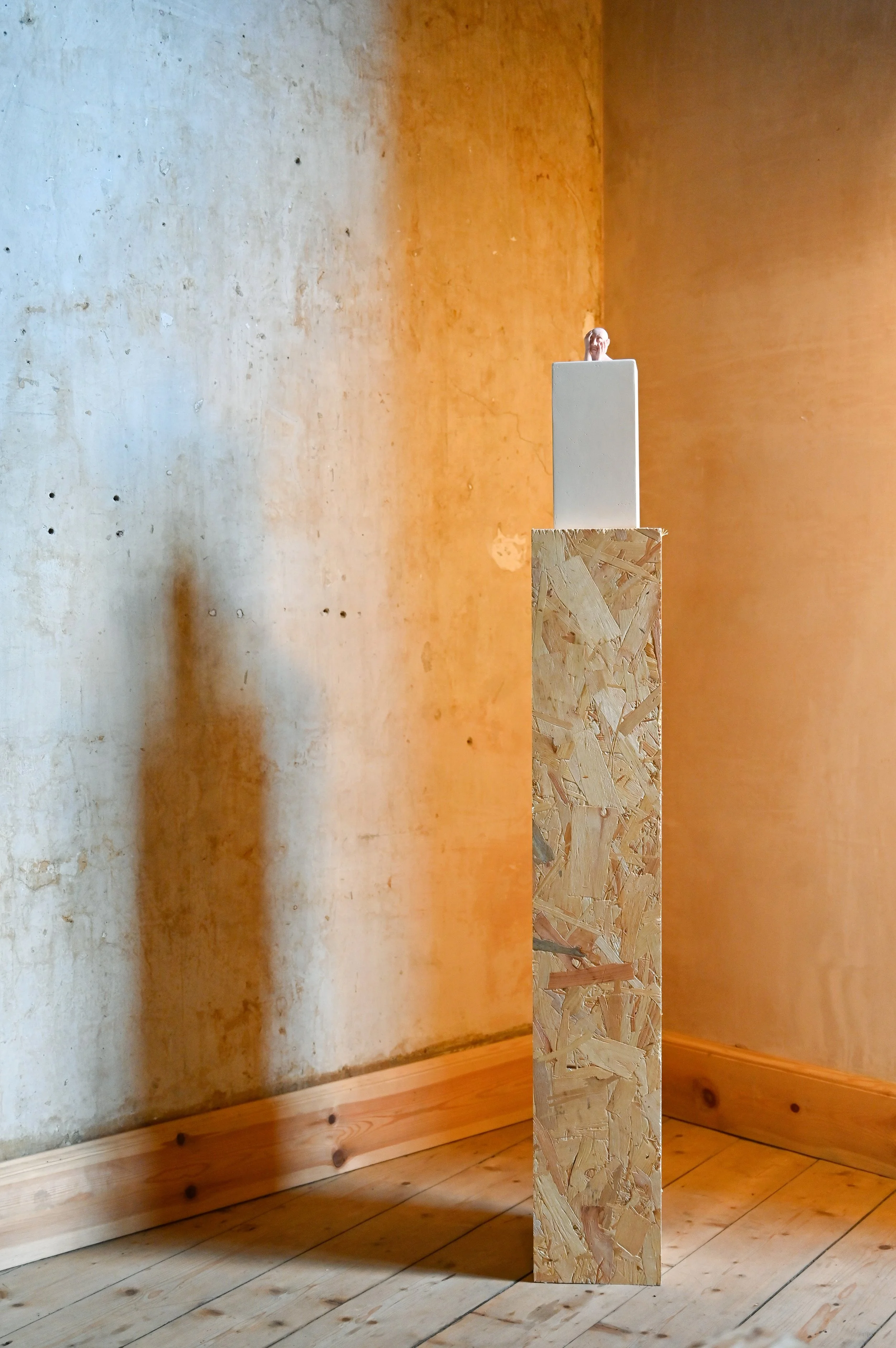 A small figurine with a pink face and white hair placed on top of a tall, rectangular wooden pedestal in a room with wooden flooring and a textured, orange-toned wall.
