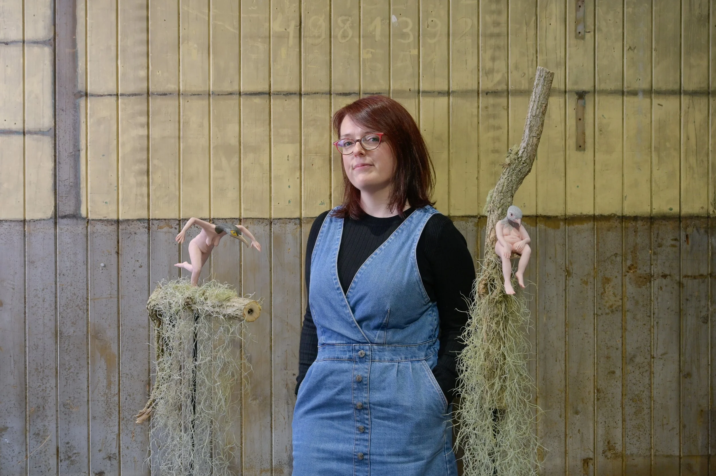 A woman with red hair and glasses wearing a black long-sleeve shirt and denim dress standing in front of a wooden wall with sculptures of human figures with bird and animal heads, connected to moss-covered tree branches.