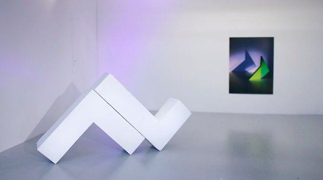 White abstract sculpture on the floor with a colorful geometric art piece on the wall behind it.