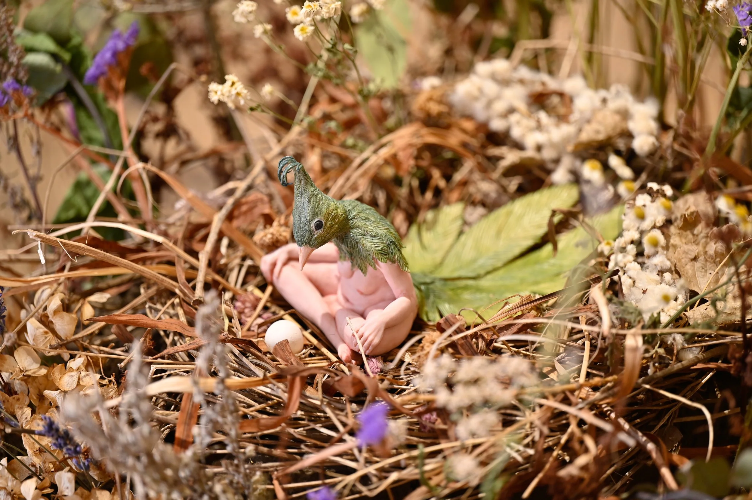 A detailed model of a human-bird hybrid with a leafy head, sitting in a nest made of twigs, leaves, and flowers, with a tiny egg nearby.