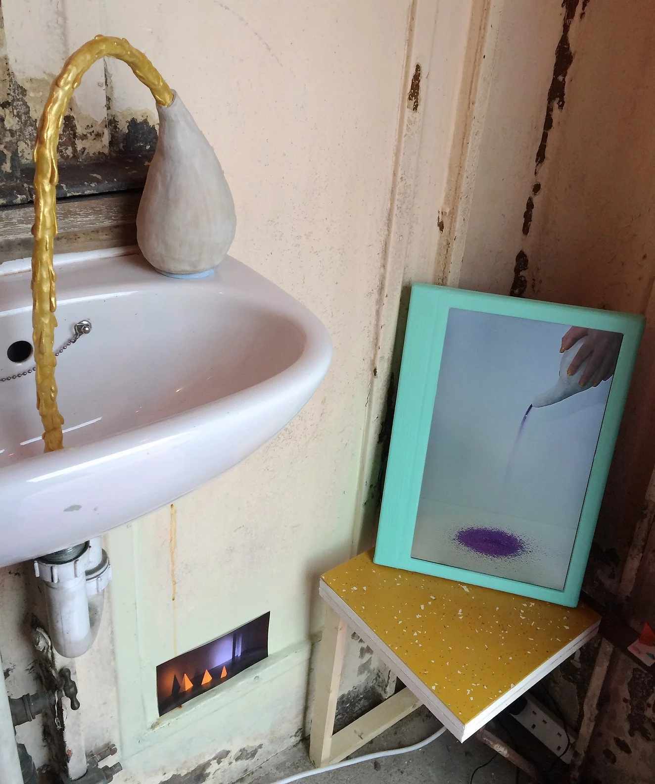 A small white sink with a ceramic vasewith yellowish water flowing, a framed picture of a hand pouring purple liquid onto a purple circle, and a yellow table with white speckles.