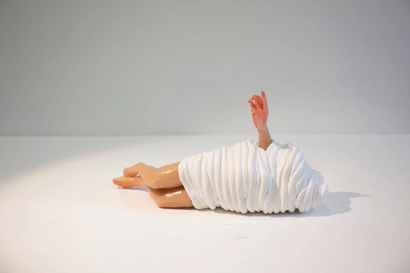 A sculpture of a figure wrapped in a white textured blanket with one hand raised, lying on a white surface against a plain background.