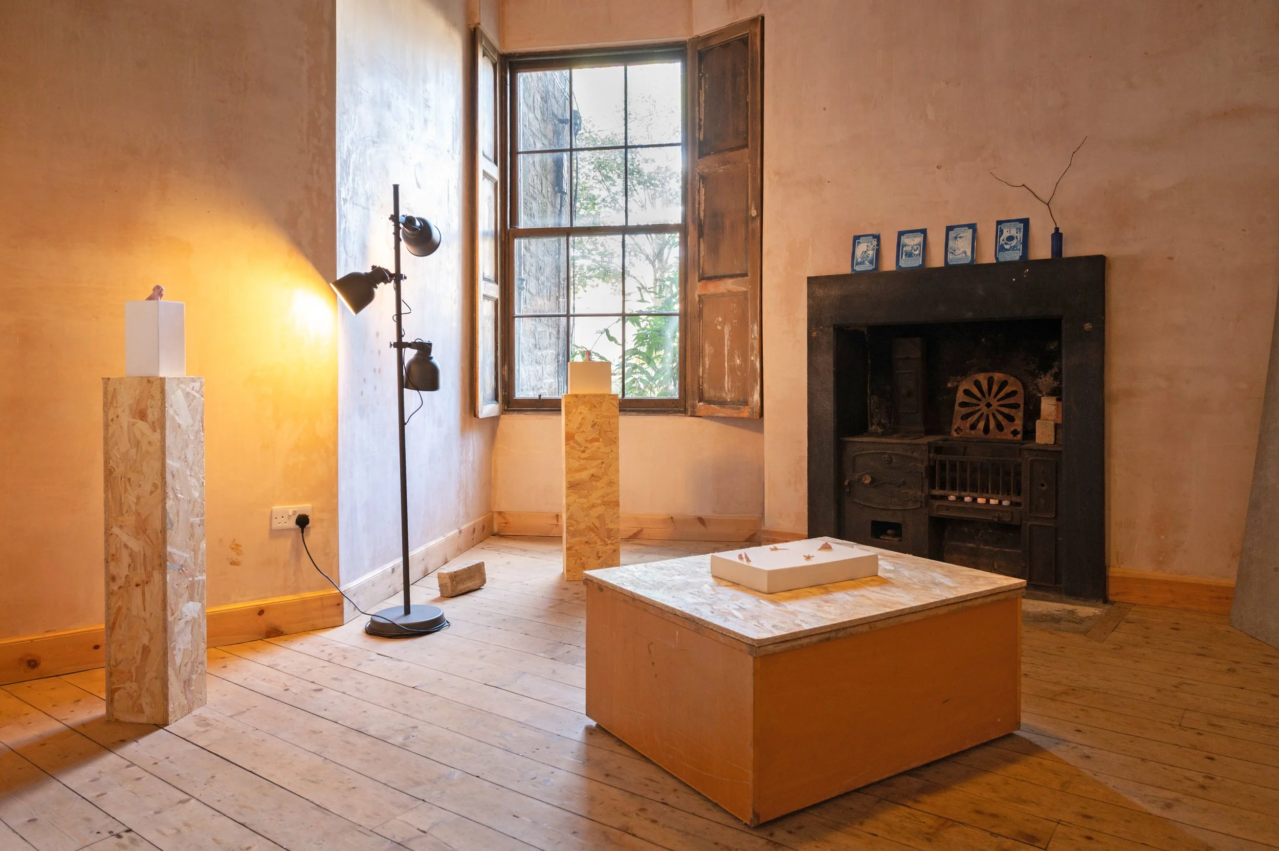 An art gallery room with a large window, a standing lamp, a pedestal with a sculpture, a low wooden table with an art piece, and a black fireplace with blue cyanotype artwork on top.