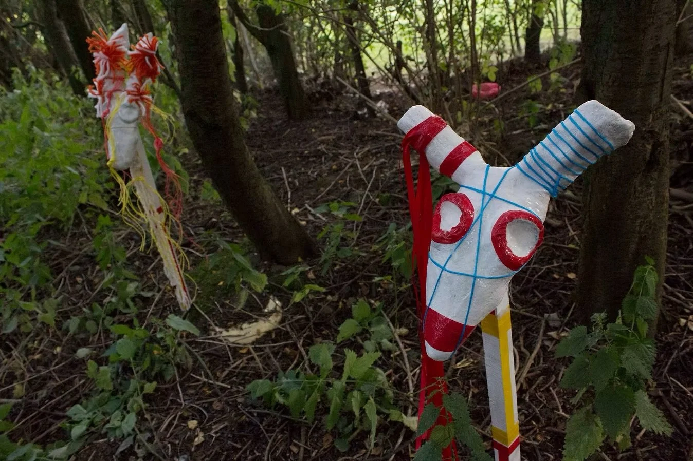 A decorated skull with red, blue, and yellow paint, tied with red and blue strings, standing amidst green bushes and trees.