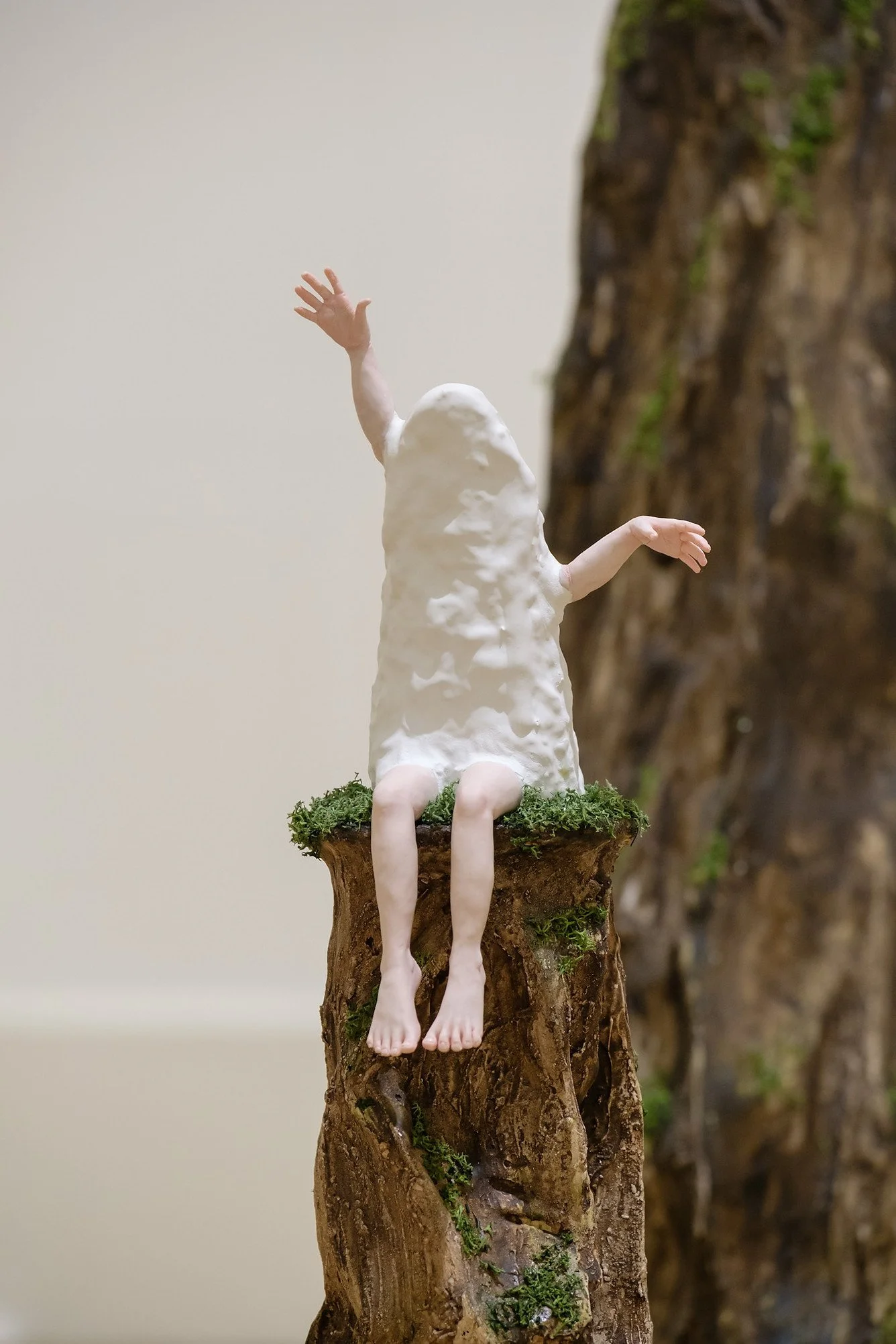 A mixed media sculpture of a human figure with a rock-like head, sitting on a tree stump with legs hanging down, and arms raised, against a plain background.