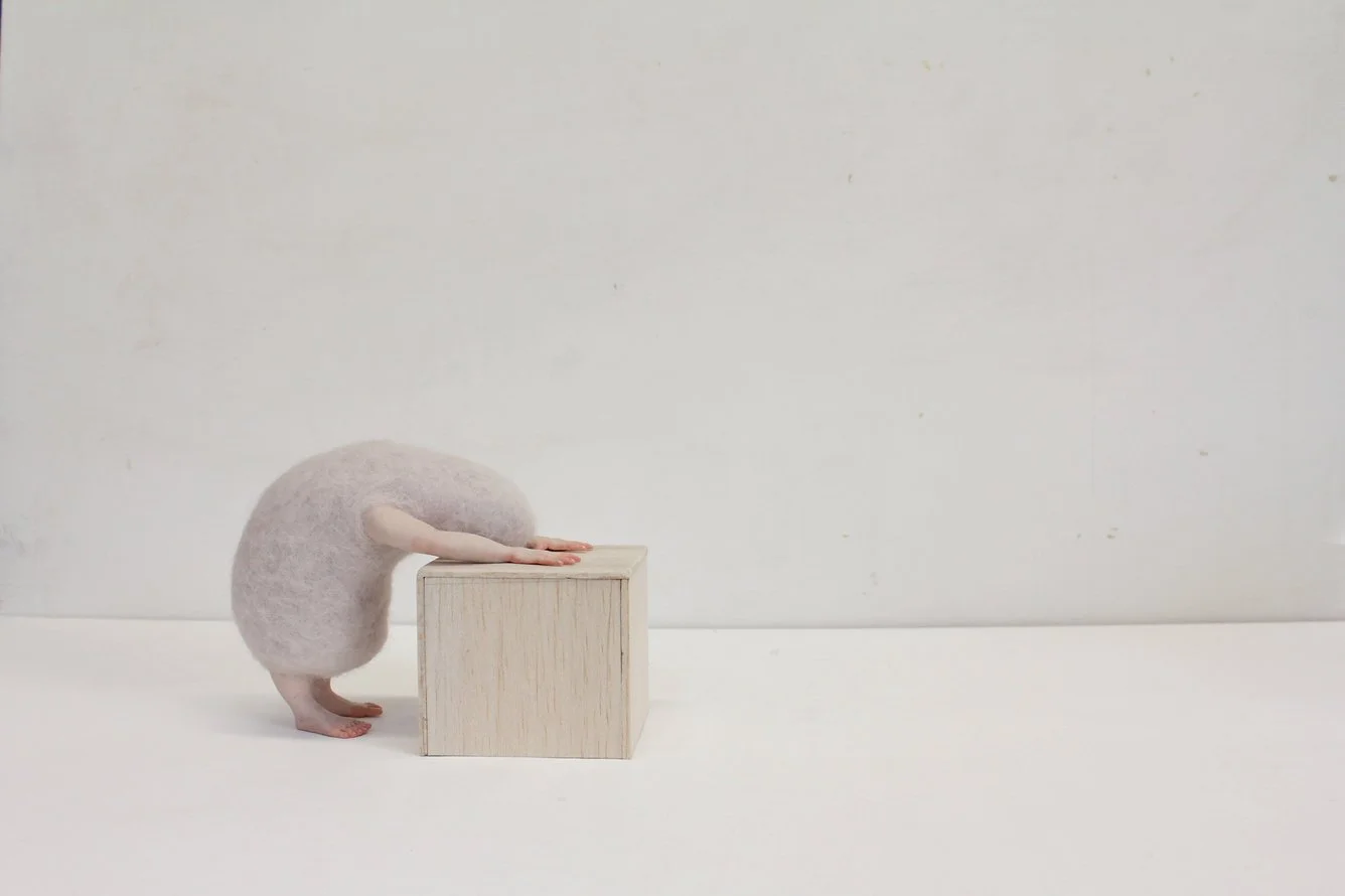 A creature with a round, fluffy body and small limbs, bent over with its forearms resting on a wooden cube, set against a plain white background.