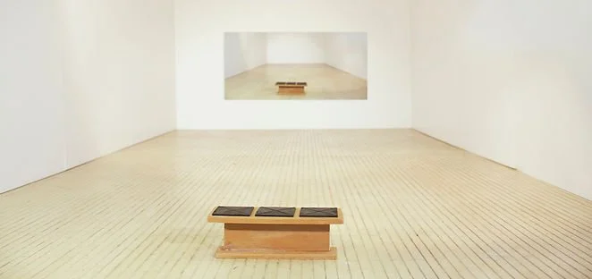 Empty art gallery with white walls, light wooden floor, and a small wooden bench in the center. There is a large mirror on the back wall reflecting the room.