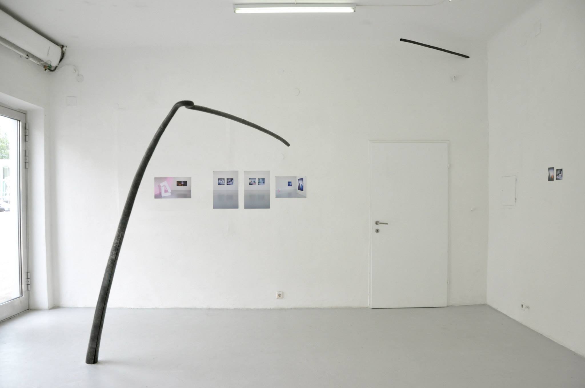 Minimalist art gallery with a white wall, featuring five small framed photographs, a white door, and a black bent pipe or sculpture leaning against the wall, with a glass door on the left.