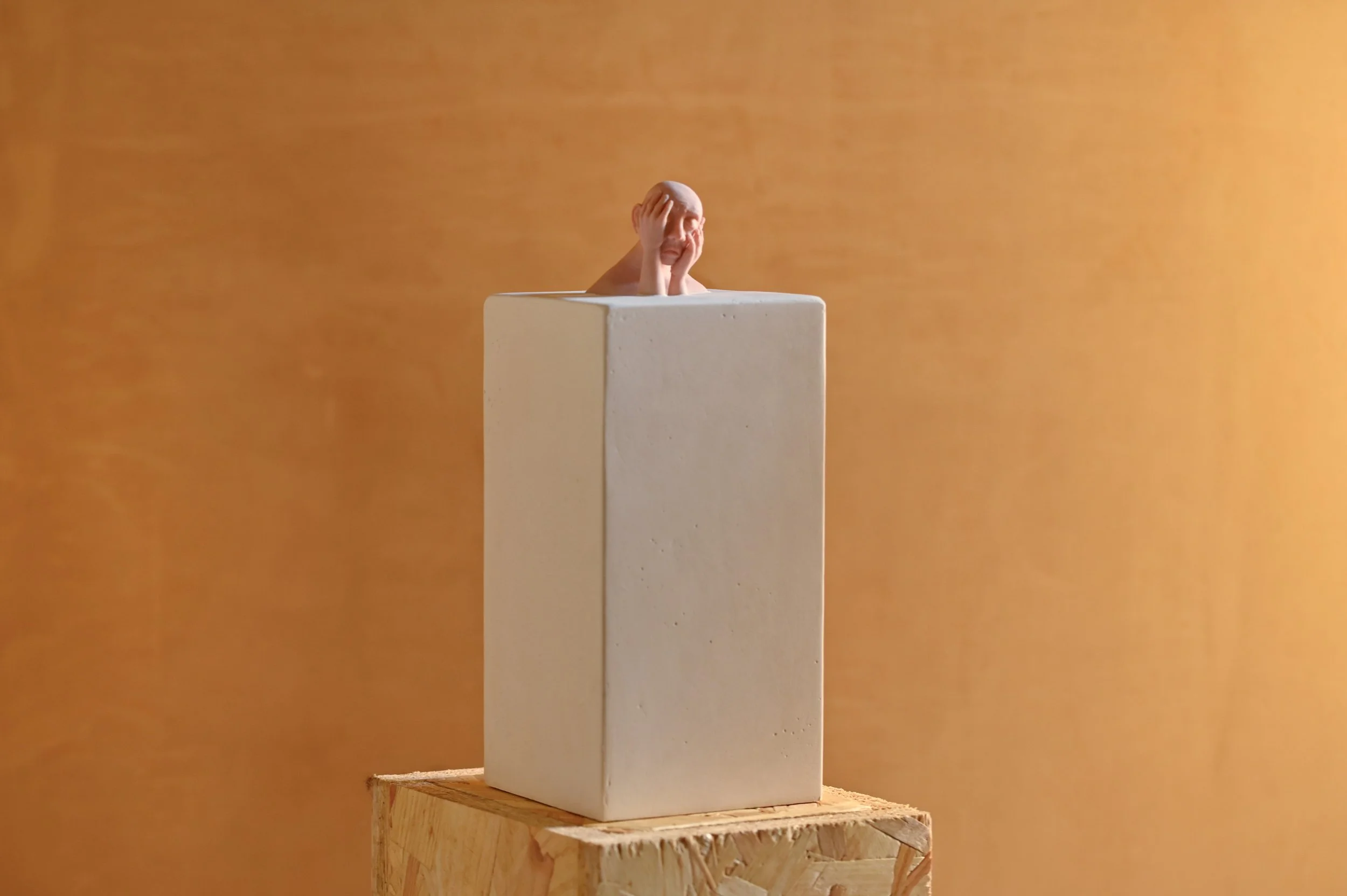 A small sculpture of a person with their hands on their face peeks out from a tall, white rectangular block, placed on a wooden surface with an orange background.