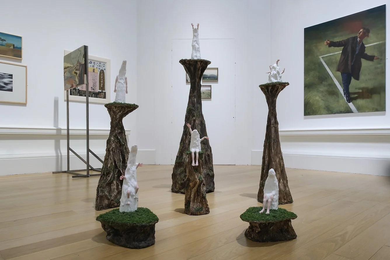 An art installation featuring several tree stumps with white plaster sculptures, some with human-like figures, in an art gallery with paintings on the walls.