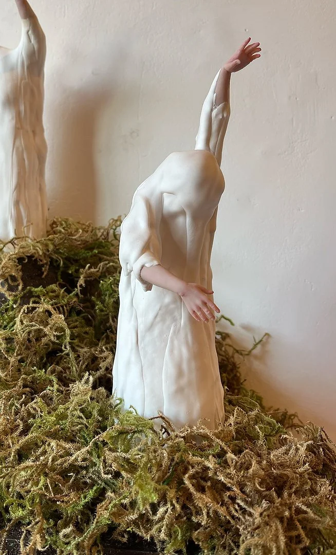 A sculpture of a human figure with a missing head, standing among dried moss or plants. The figure has one arm raised above the head and another across the body, in a contemplative pose.