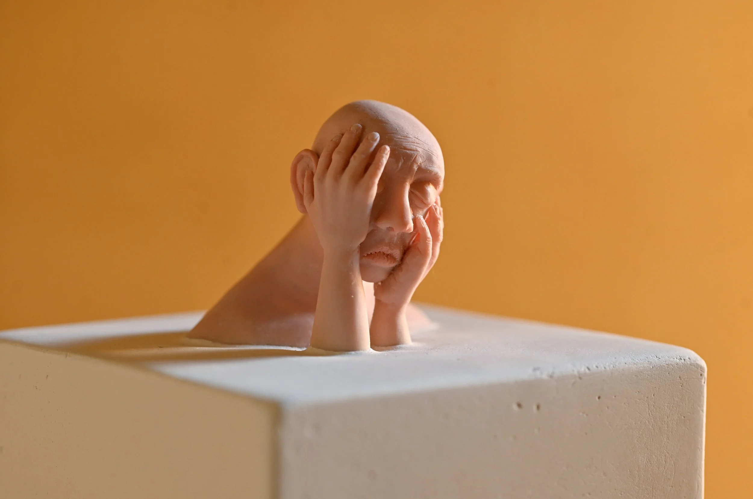 Sculpture of a man with a bald head, covering his face with his hands, placed on a white pedestal against a warm orange background.
