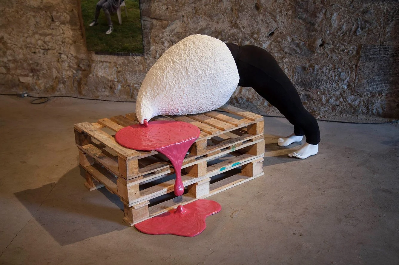 An art installation of a person with a large, white, textured head, bent over with their hands and feet on the ground, appearing to inspect or interact with a wooden pallet. The person is wearing black pants and white socks. The background has a ston