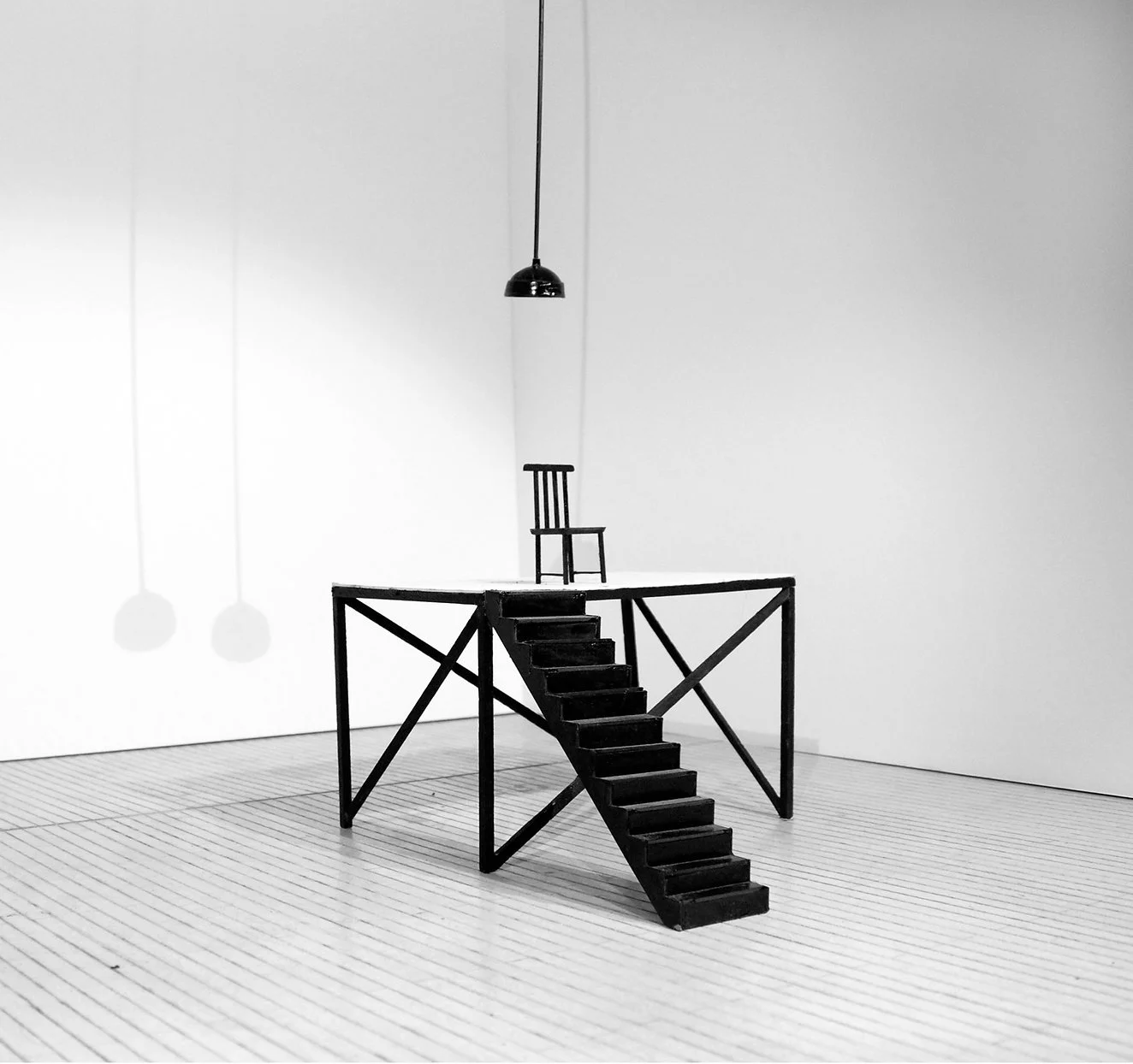 A black mini staircase leading to a platform with a small black chair on top, set against a plain white wall in a minimalistic room, with shadows cast on the wall by hanging lamps.