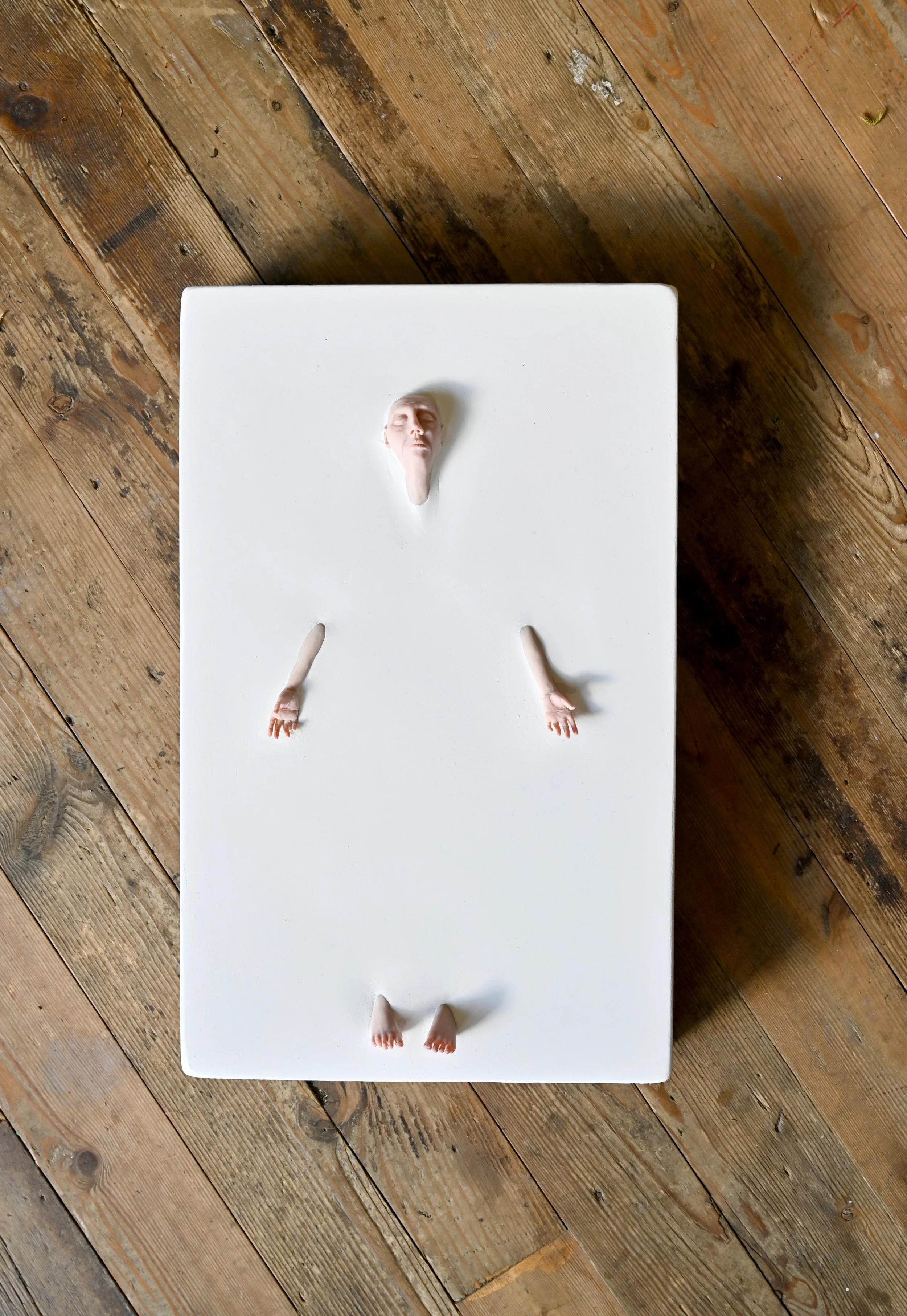 An art piece featuring a human face and limbs emerging from a white rectangular surface, with the face at the top, hands in the middle, and feet at the bottom, placed on a wooden floor.