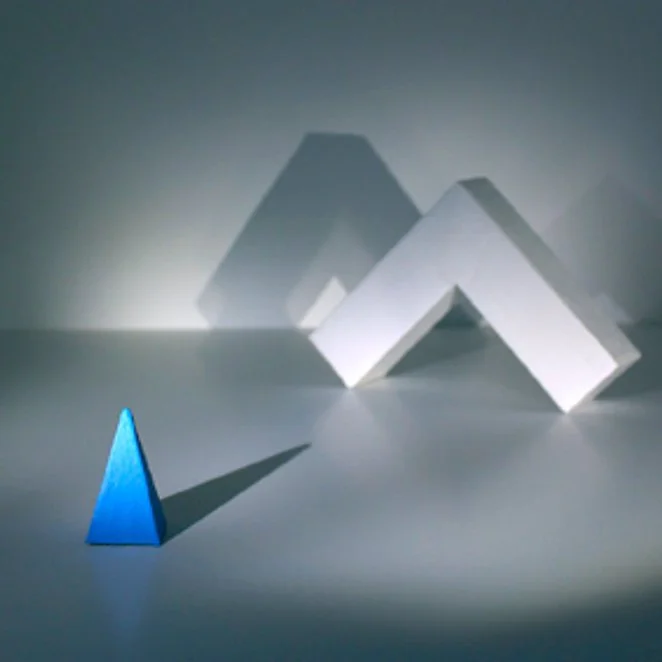 A blue triangular prism and three white rectangular blocks casting shadows on a gray surface, creating an abstract geometric arrangement.
