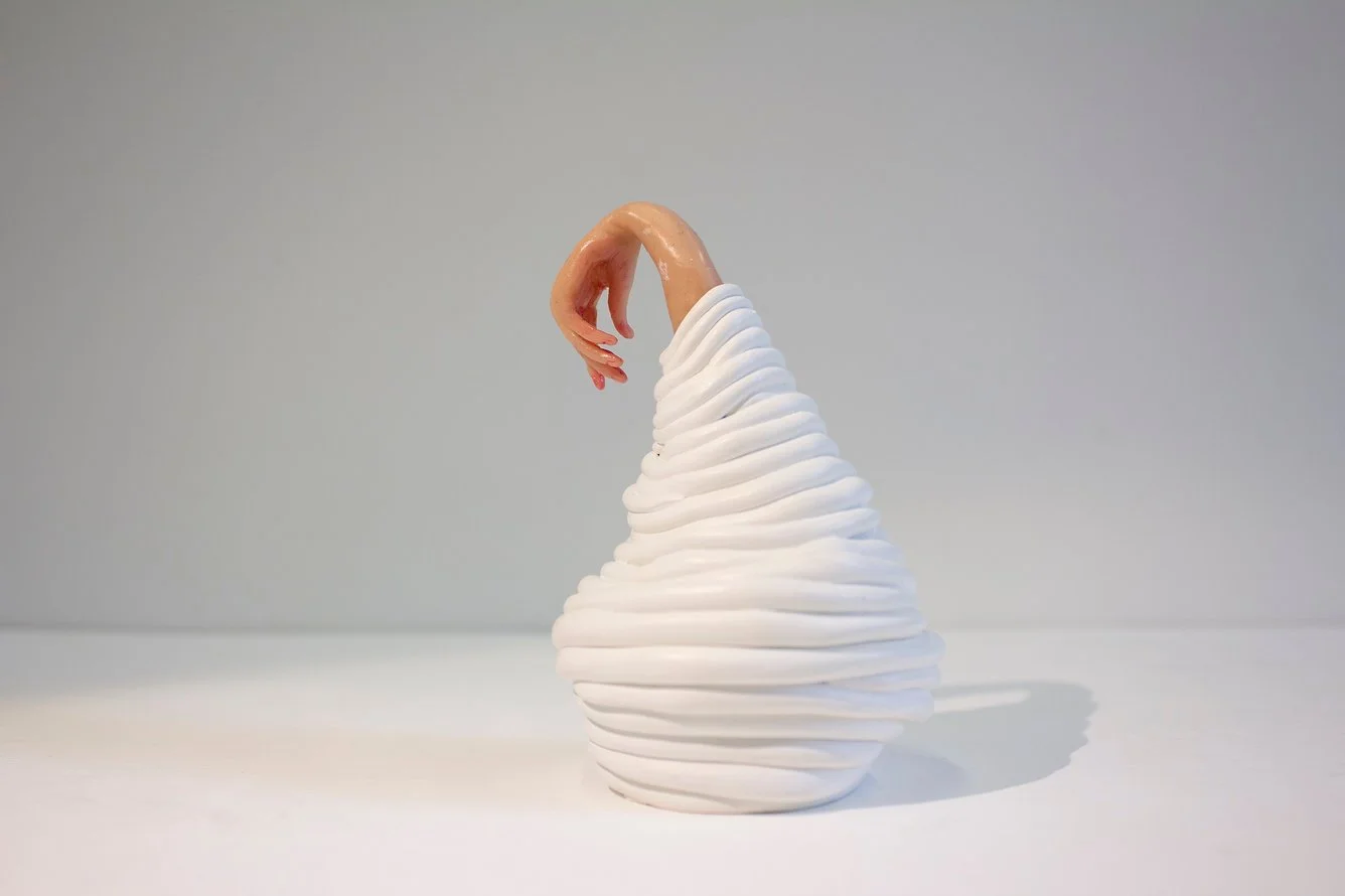 A sculpture of a hand emerging from a swirl of white material resembling whipped cream or meringue, set against a plain gray background.