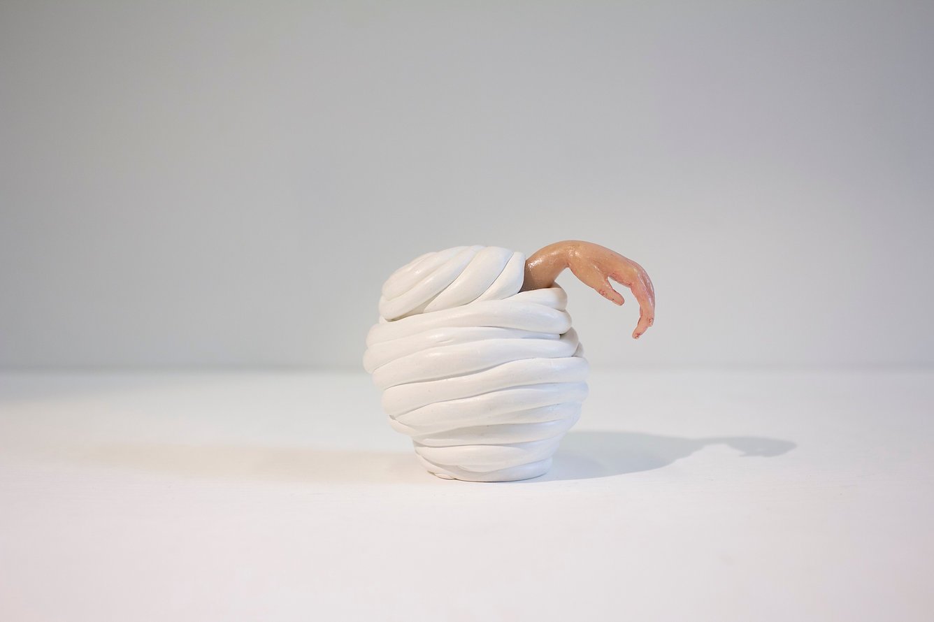 An artistic sculpture of a swirl of white material resembling whipped cream or meringue with a human hand emerging from the top, set against a plain background.