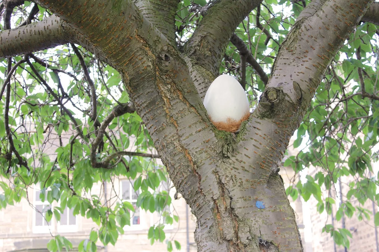 A white artificial bird egg resting in a tree branch with green leaves.