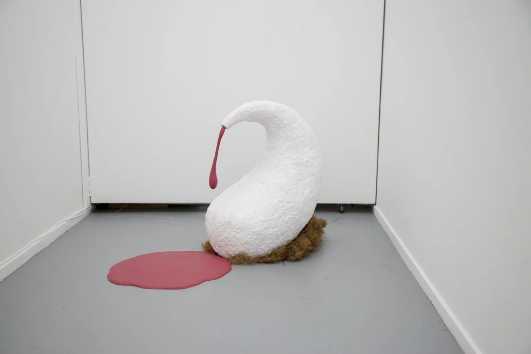 A sculpture of a swan with a red beak, with pink paint spilled on the floor and puddled underneath, on a light gray floor in a white corner.