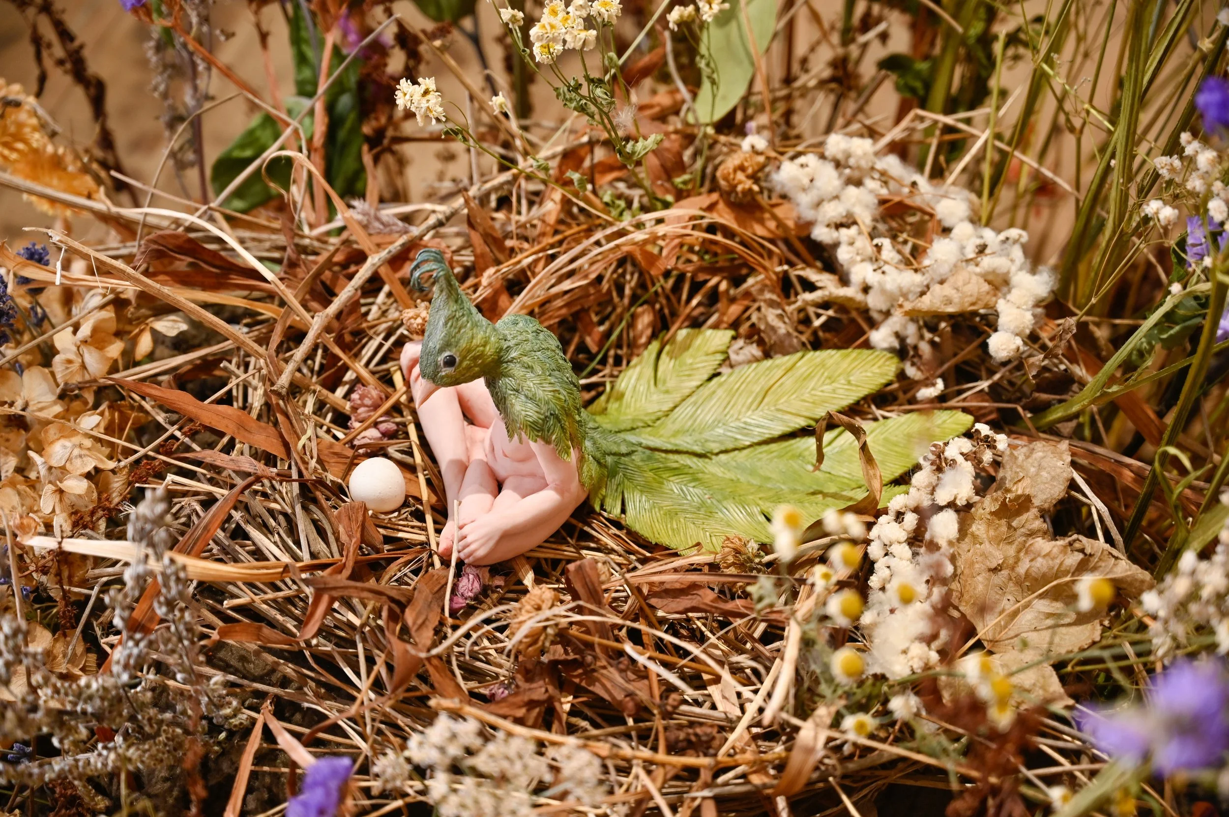 A detailed miniature scene of a human-bird hybrid with a leaf tail, surrounded in a nest of dried leaves, flowers, and natural elements.