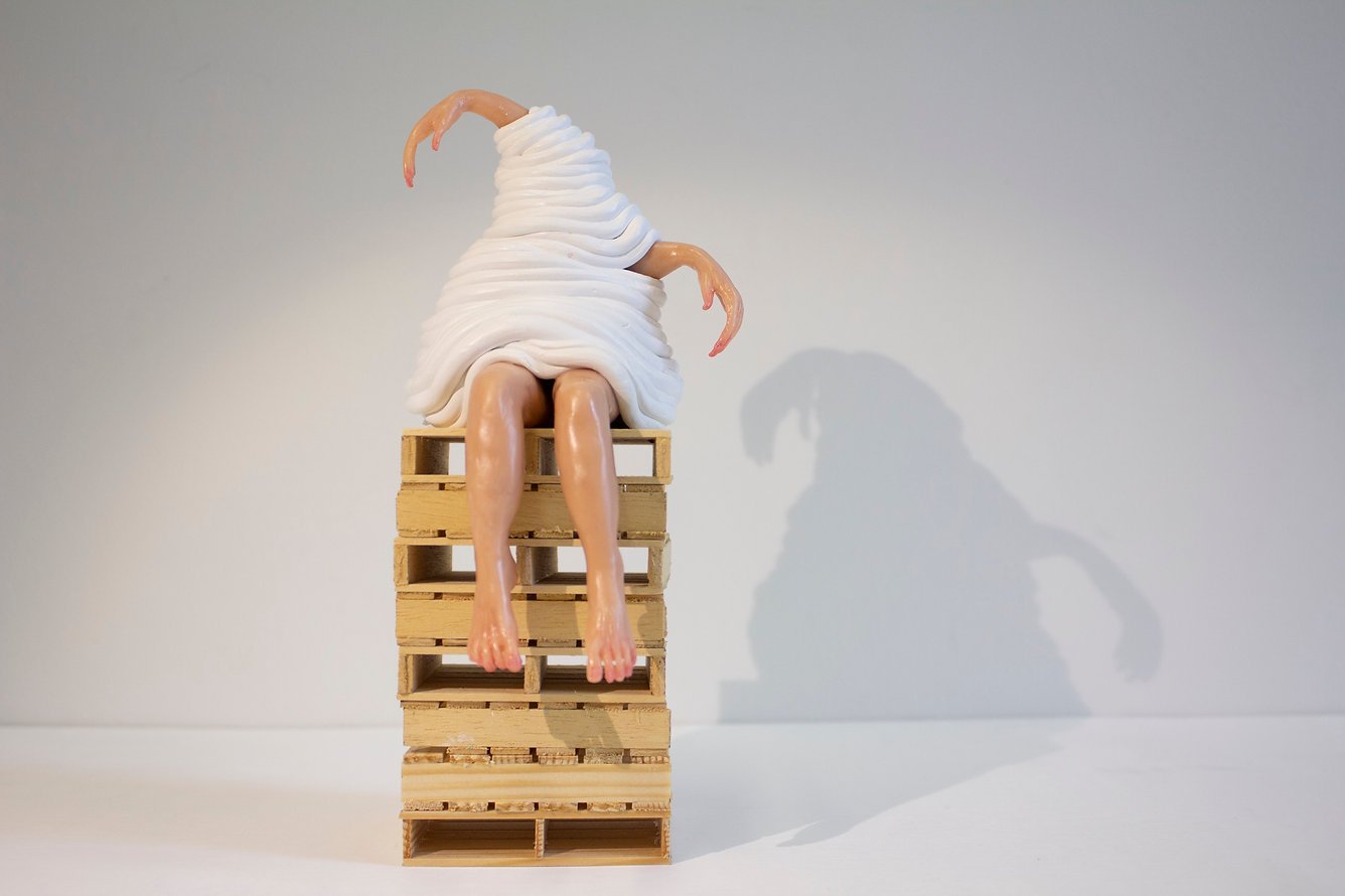 A sculpture of a person with a swirl of white material covering their upper body, sitting on a wooden pallet with their legs hanging over the edge. The sculpture has arms with shrimp-like hands and feet, and casts a shadow on the plain wall behind.