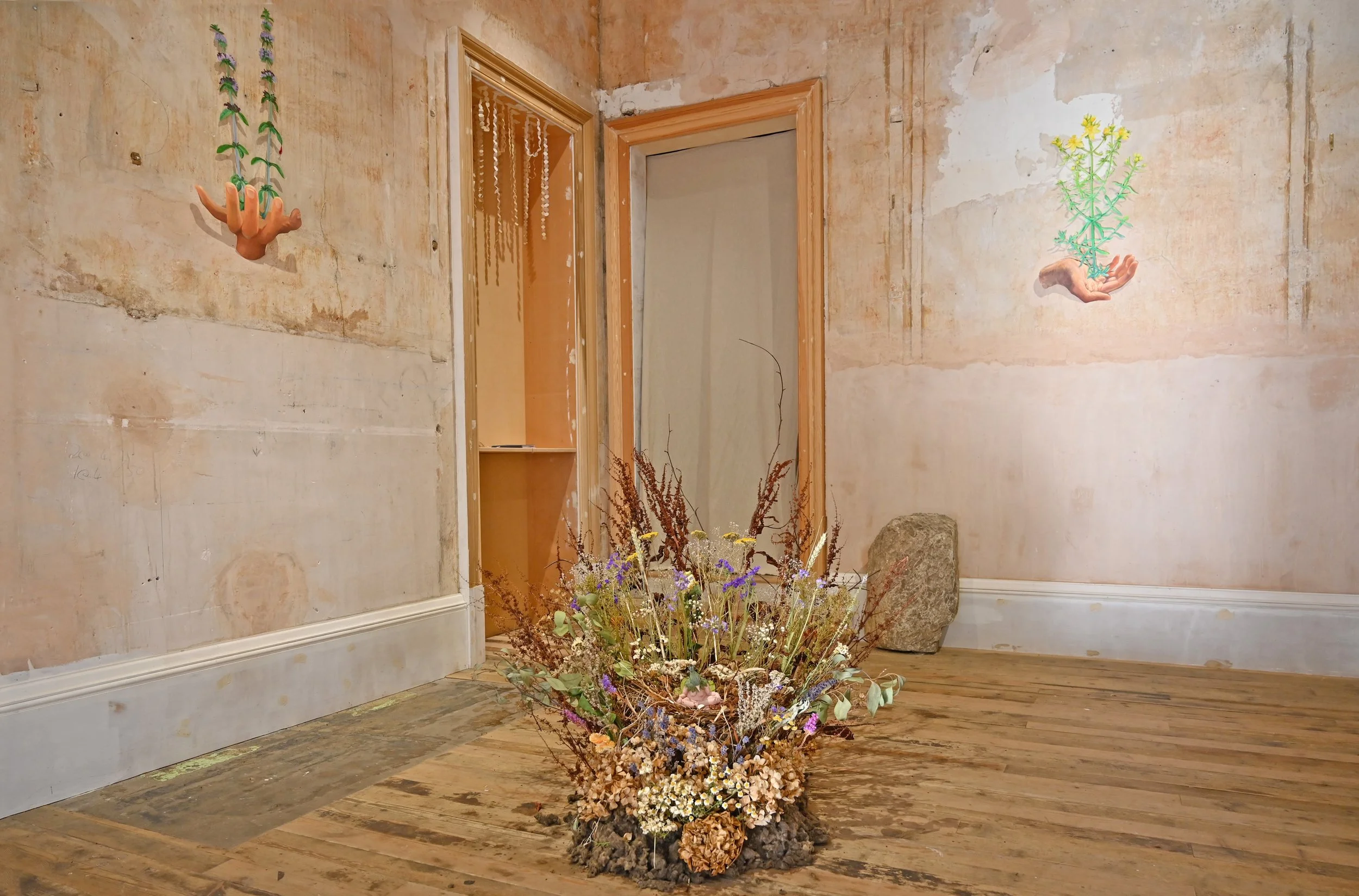 An indoor art installation featuring a beige, weathered wall with two hanging plant-themed sculptures of hands holding plants, a large flower arrangement on a wooden floor, and a door with a beaded curtain.