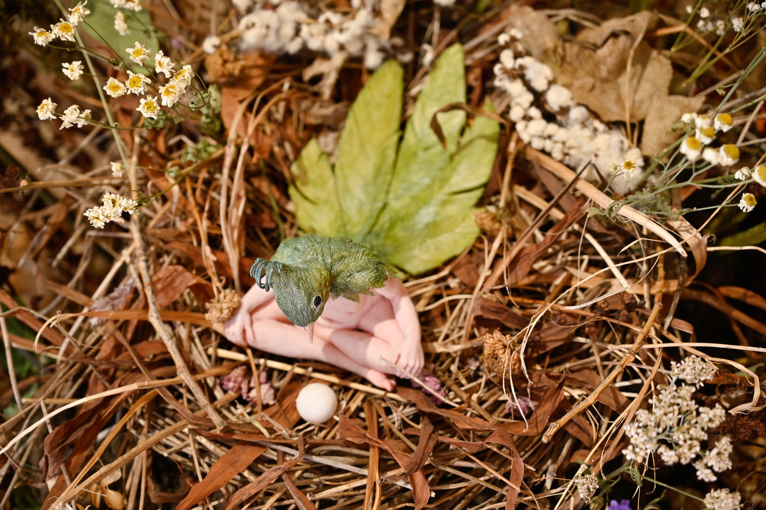 A small green hybrid human-bird figure sitting in a nest made of dried grass and surrounded by small white flowers with yellow centers.